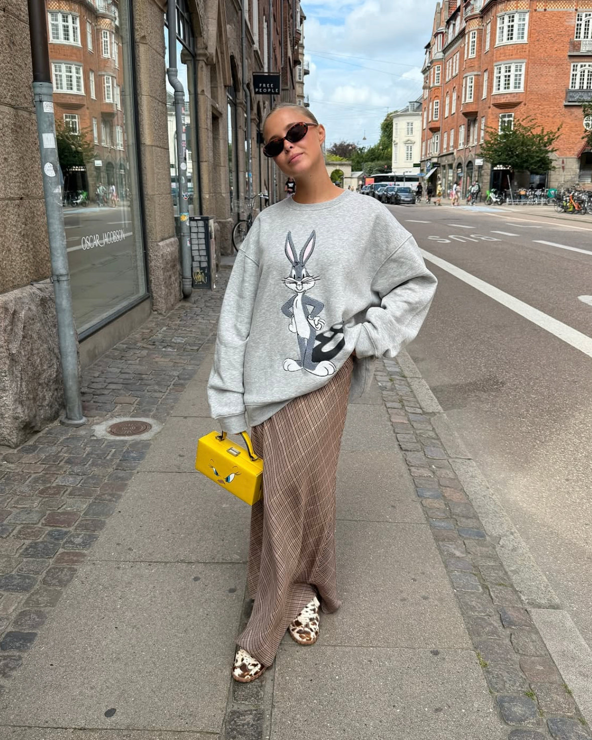 Person wearing a gray sweater with a graphic design, brown pants, and a yellow bag on a city street.