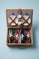 A wicker picnic basket with its lid open, styled with a picnic set and four vintage sneakers inside. The basket contains two white plates with brown leather straps, two striped ceramic cups, and four sneakers in green, burgundy, navy, and brown.